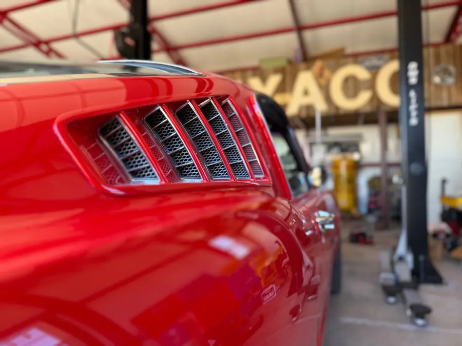 Close-up of the chrome quarter-panel louvers on a red Mustang fastback, with the shop's Texaco sign blurred behind